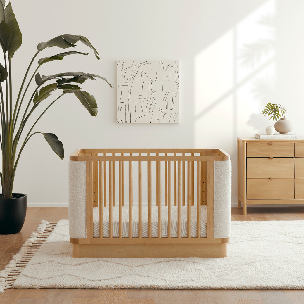 Front view of Babyletto Bondi Boucle 4-in-1 Convertible Crib next to a plant and dresser in -- Color_Honey with Ivory Boucle
