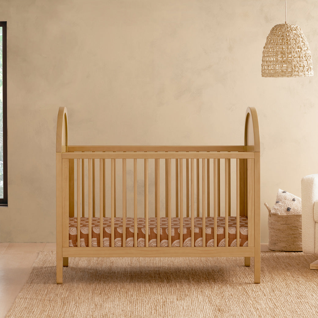 Front view of Babyletto Bondi Cane 3-in-1 Convertible Crib next to a basket and recliner  in -- Color_Honey with Natural Cane