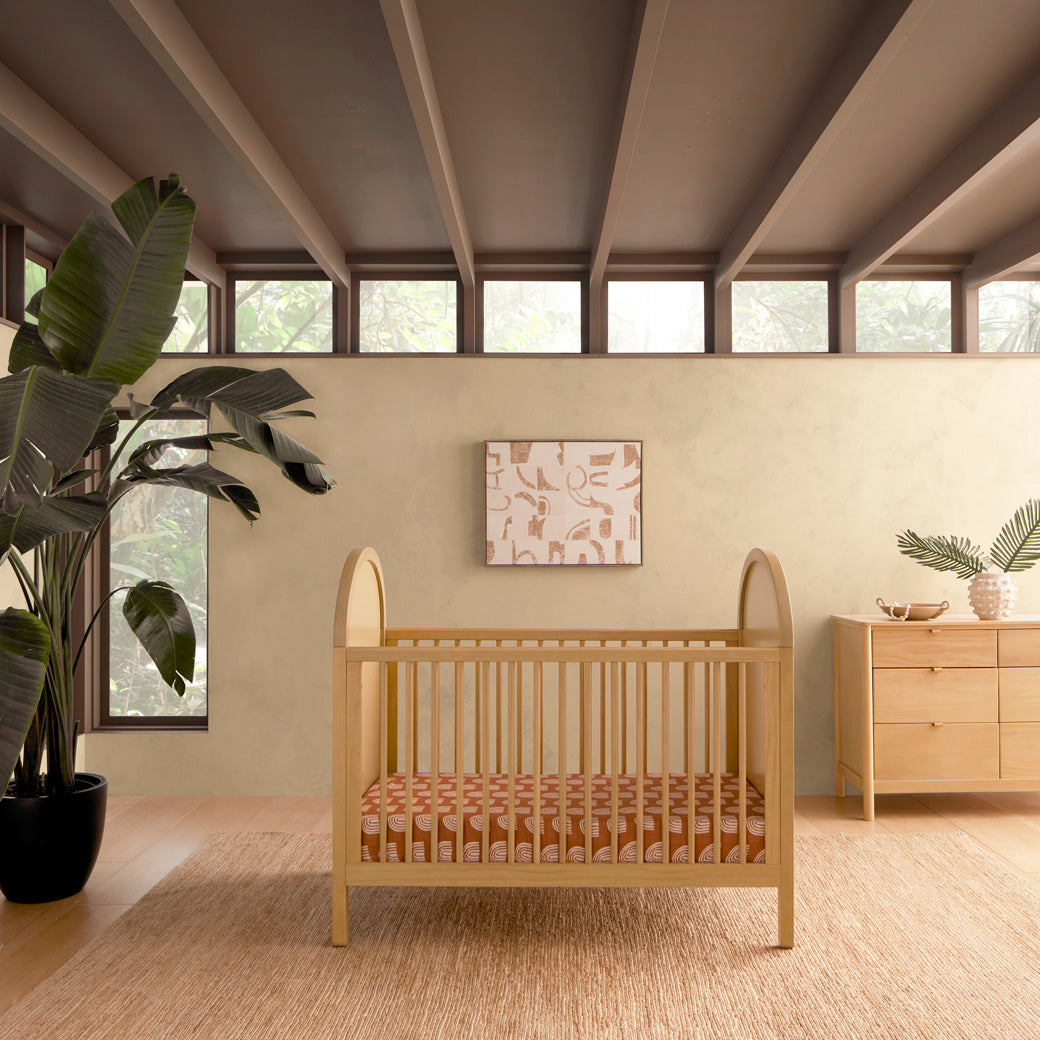 Front view of Babyletto Bondi Cane 3-in-1 Convertible Crib next to a plant and a dresser in -- Color_Honey with Natural Cane