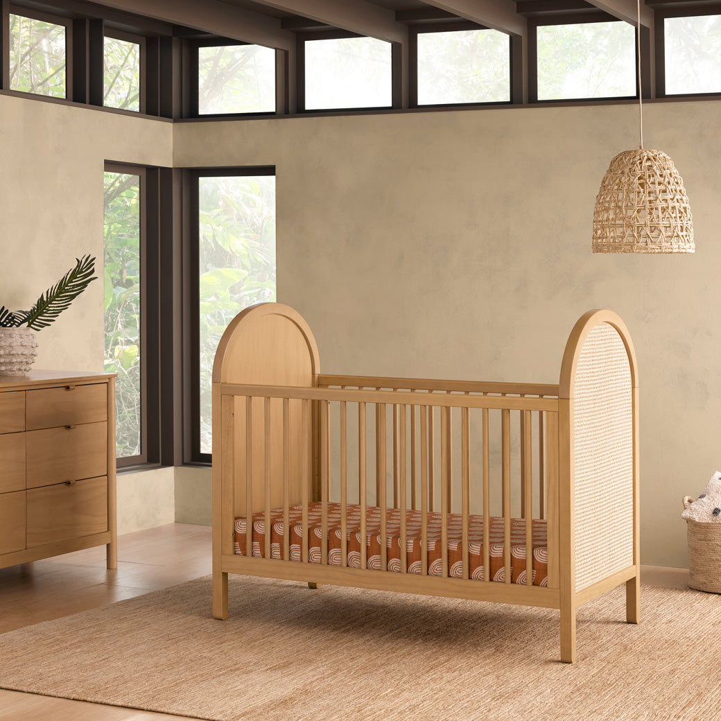 Babyletto Bondi Cane 3-in-1 Convertible Crib under a lamp and next to a dresser in -- Color_Honey with Natural Cane