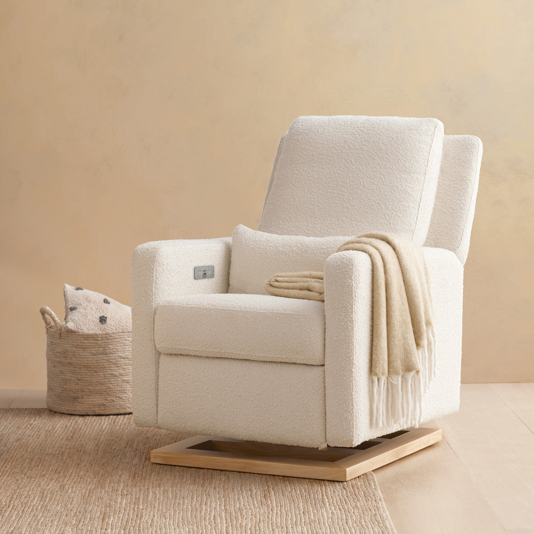 Babyletto Sigi Electronic Glider Recliner  next to a basket and with a blanket on the seat in -- Color_Ivory Boucle