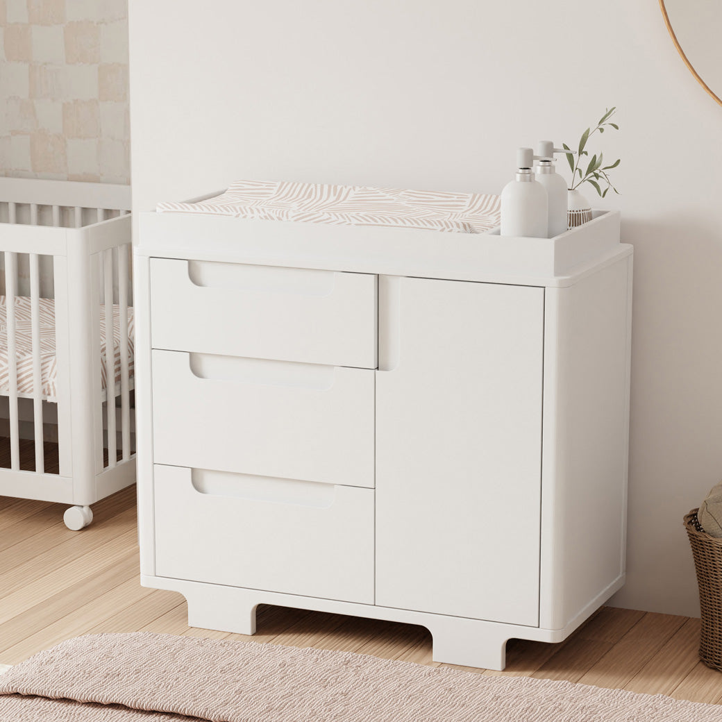 Babyletto Yuzu 3-Drawer Changer Dresser  next to a crib and basket in -- Color_White