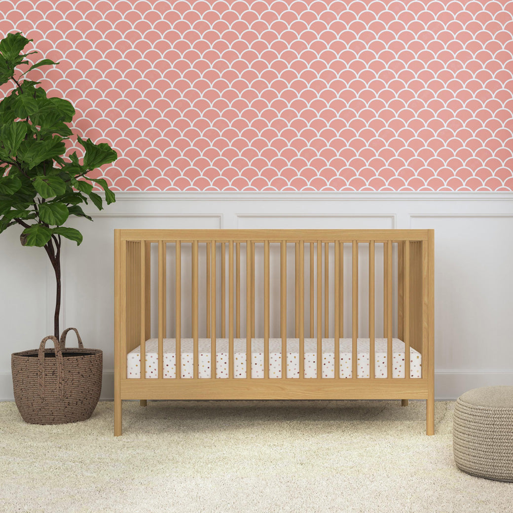 Front view of DaVinci Birdie 3-in-1 Convertible Crib next to a plant and ottoman  in -- Color_Honey