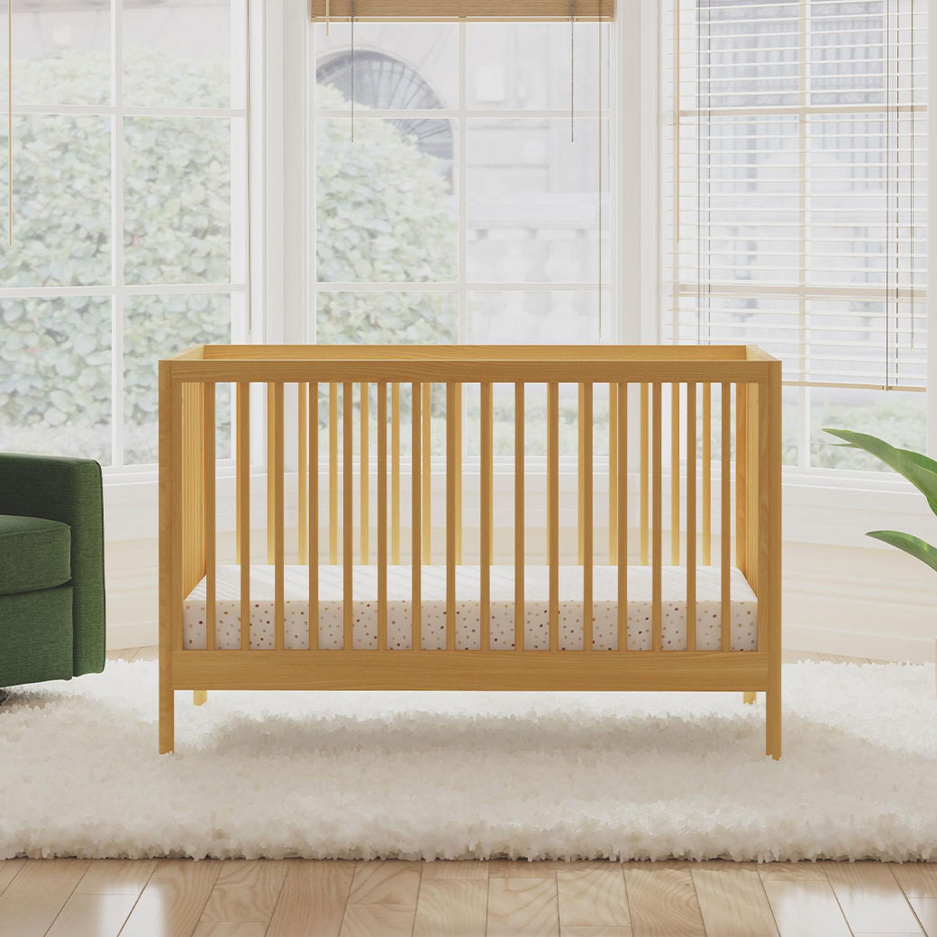 Front view of DaVinci Birdie 3-in-1 Convertible Crib next to a recliner  in -- Color_Honey