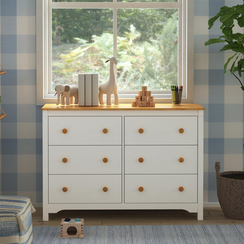 Front view of DaVinci Shea 6-Drawer Dresser under a window next to a plant  in -- Color_Warm White/Honey