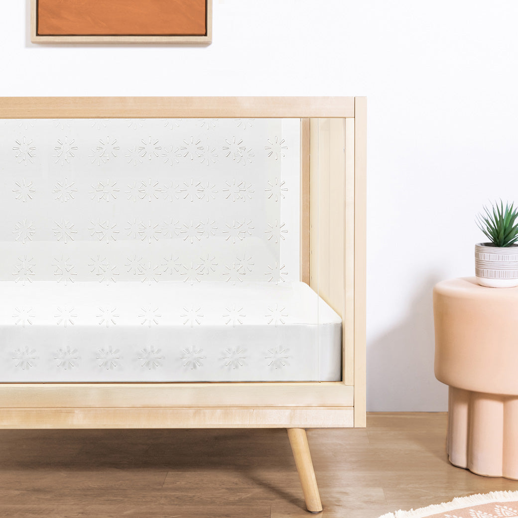 Closeup view of Ubabub Nifty Clear 3-in-1 Crib next to a plant in -- Color_Natural Birch