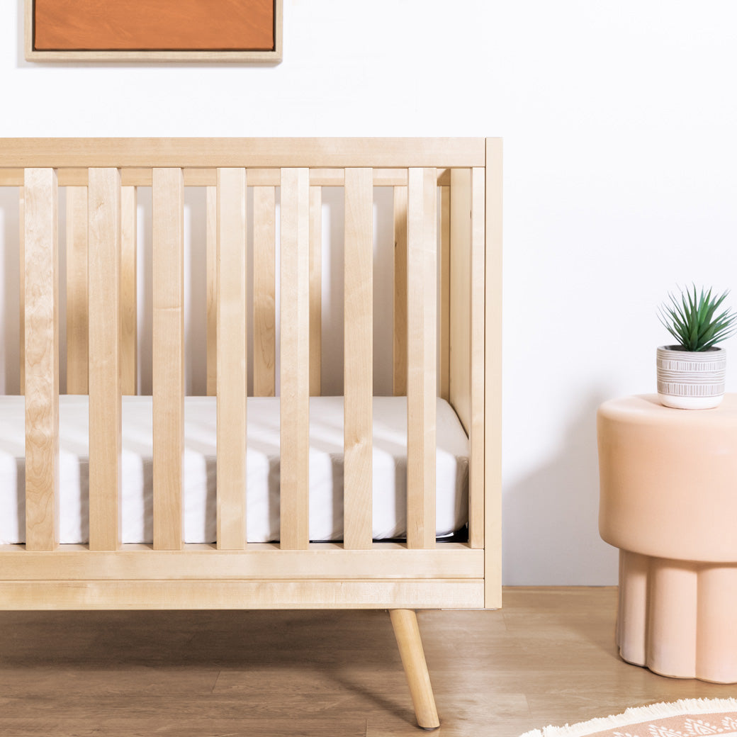 Closeyp of Ubabub Nifty Timber 3-In-1 Crib next to a plant in -- Color_Natural Birch
