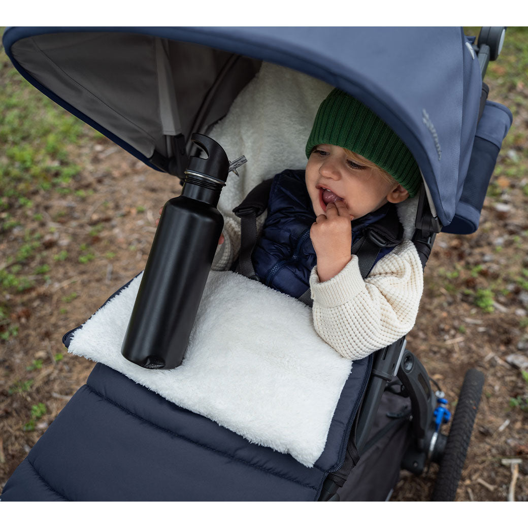 A baby in the Uppababy CozyGanoosh on a stroller holding a water bottle in -- Color_Noa