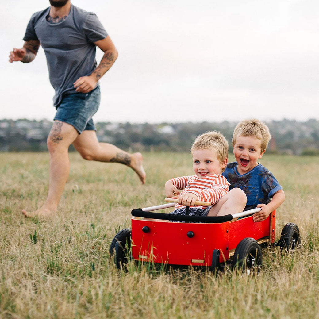 Dad running beside two boys playing in the Wishbone Wagon in Red