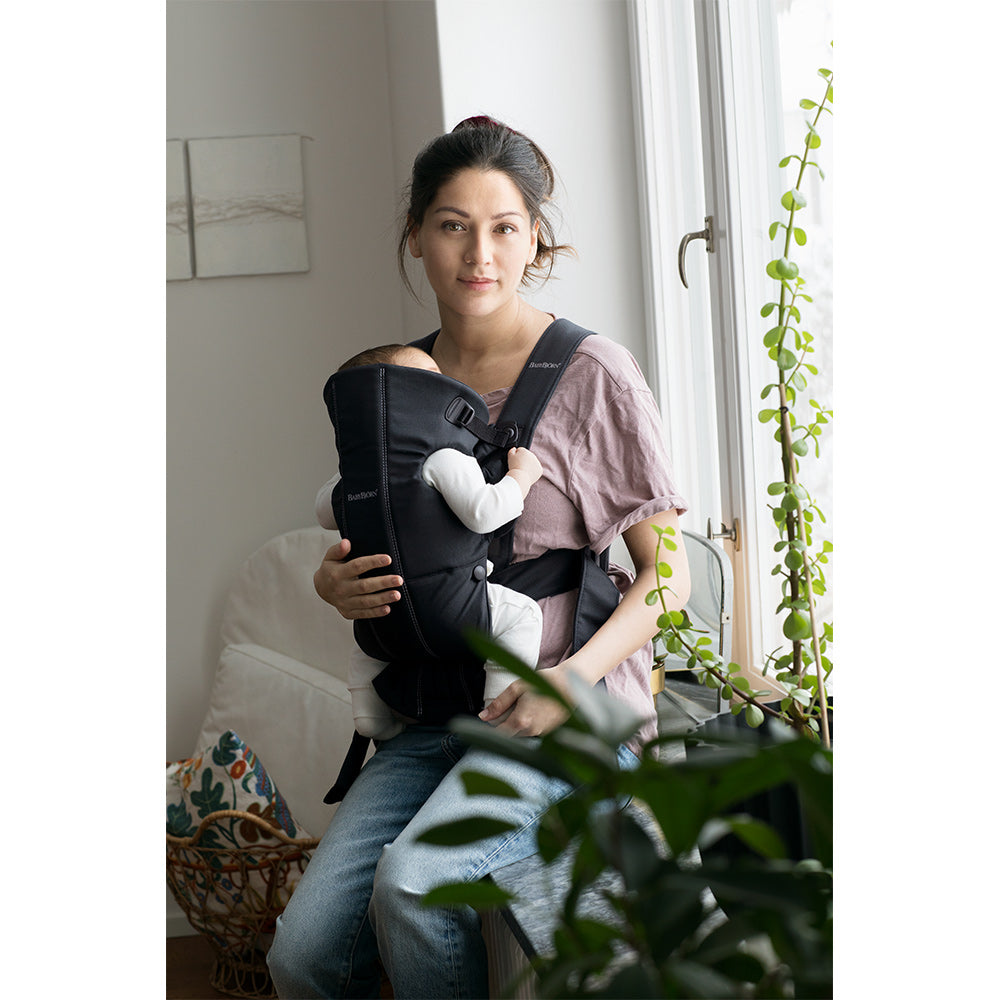 Mom sitting by a window carrying baby in BABYBJÖRN Baby Carrier Mini in -- Color_Black Woven