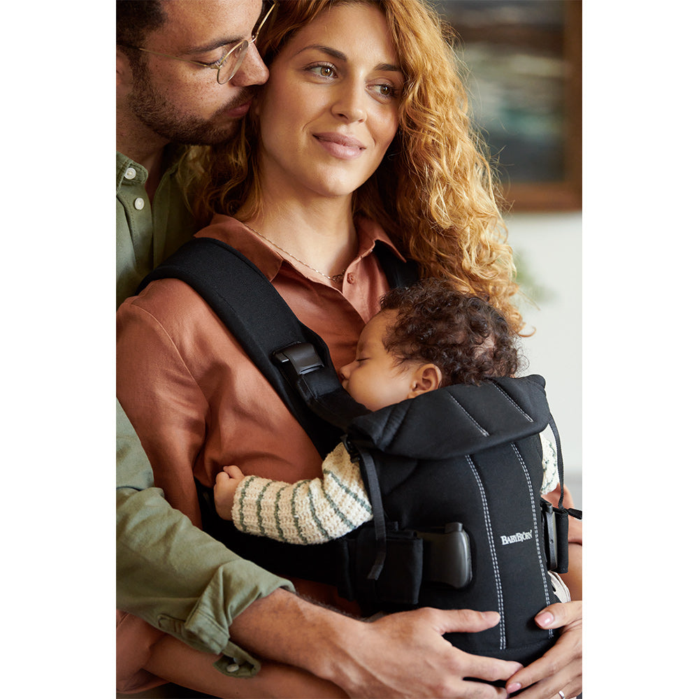 Dad has his arms wrapped around moms stomach trying to support the BABYBJÖRN Baby Carrier One in -- Color_Black Woven Mix