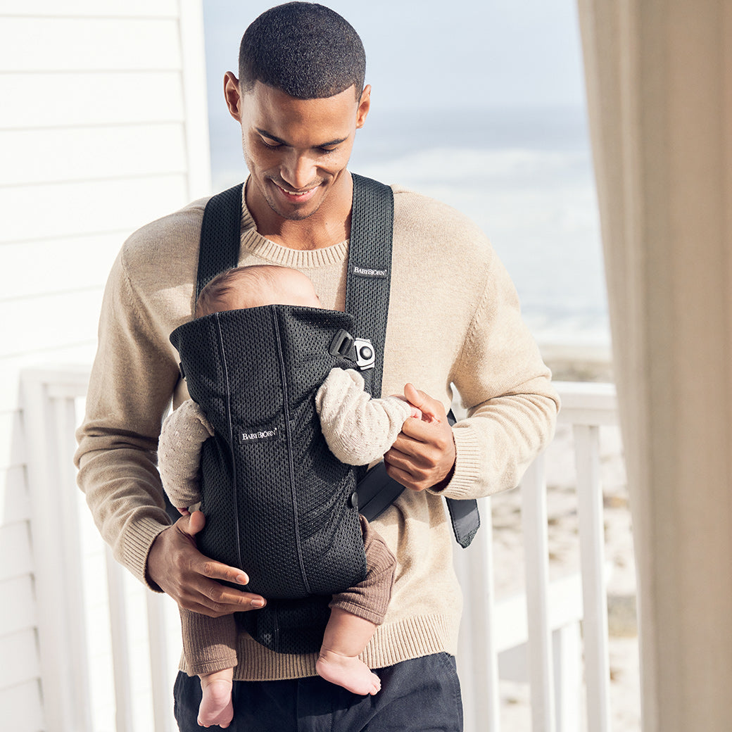 Dad standing on balcony holding baby in BABYBJÖRN Baby Carrier Mini with top part down in -- Color_Black 3D Mesh