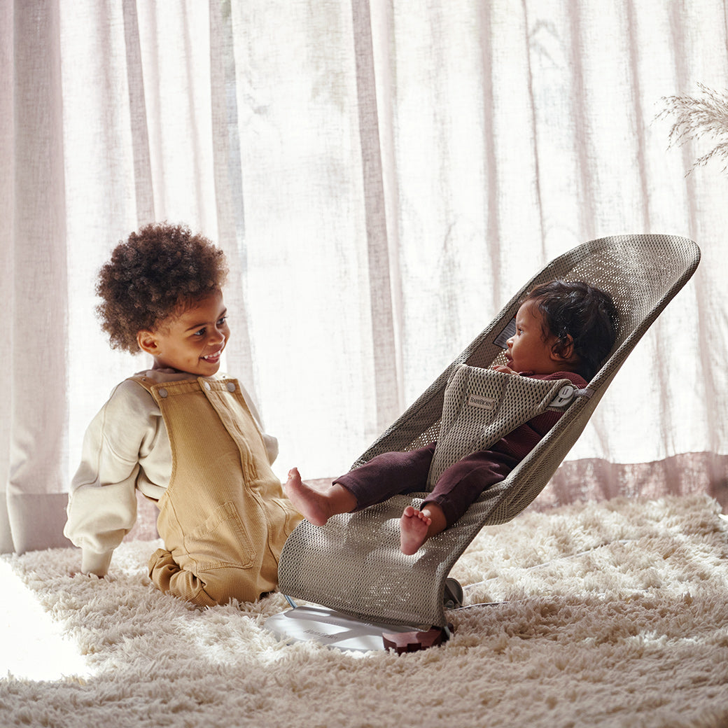 Toddler next to baby in BABYBJÖRN Bouncer Bliss on a fluffy rug in -- Color_Beige Grey Mesh
