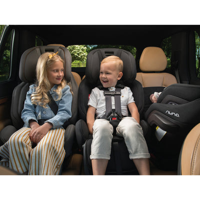 Two children sitting in the back seat of the car in their Nuna EXEC Car Seat in Color_Riveted — All