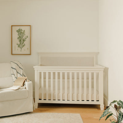 Darlington 4-in-1 Convertible Crib in Warm White next to a picture and a recliner