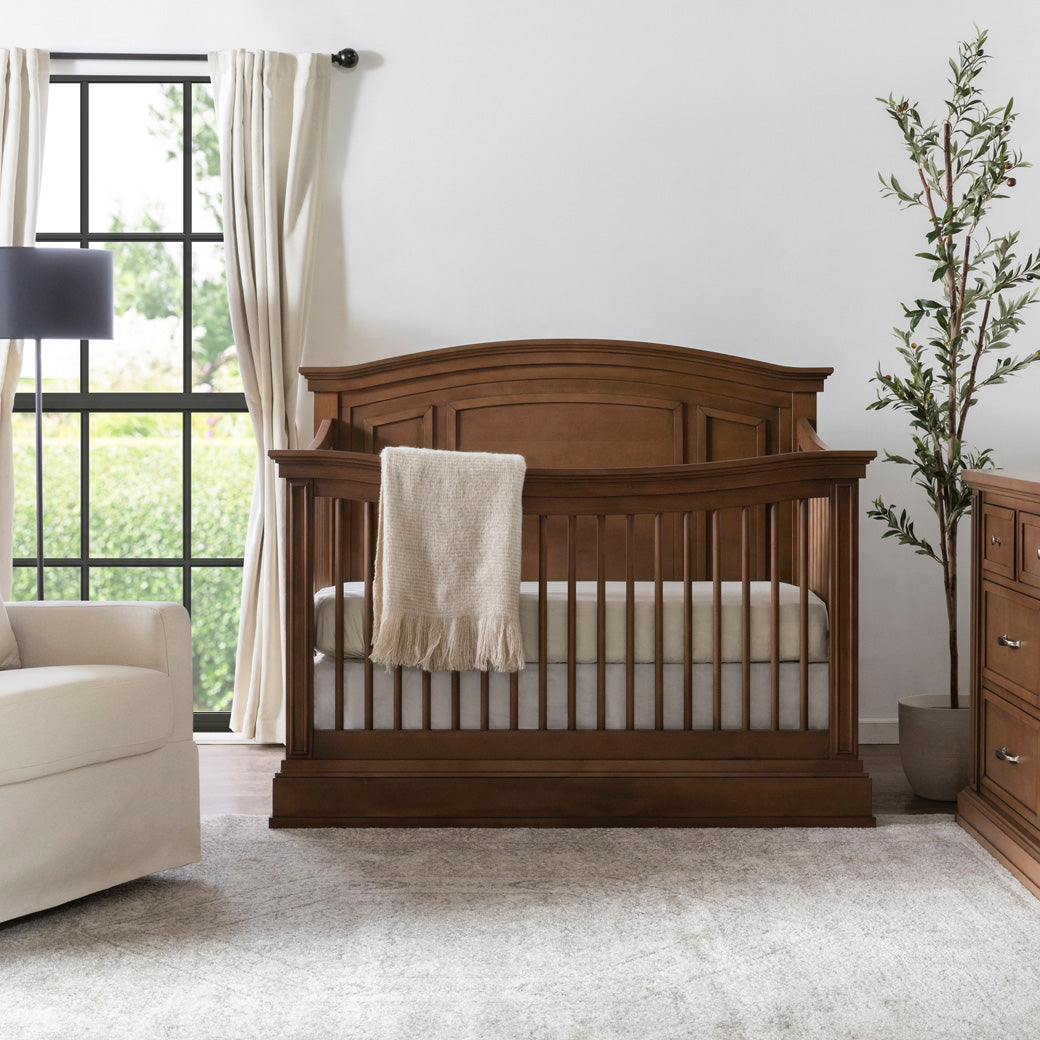 Namesake's Durham 4-in-1 Convertible Crib in a cozy room next to a recliner and dresser  in -- Color_Derby Brown
