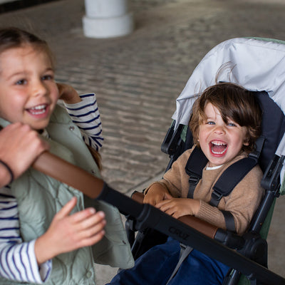 A child smiling next to a toddler sitting in the UPPAbaby Cruz V2 Stroller in -- Color_Gwen