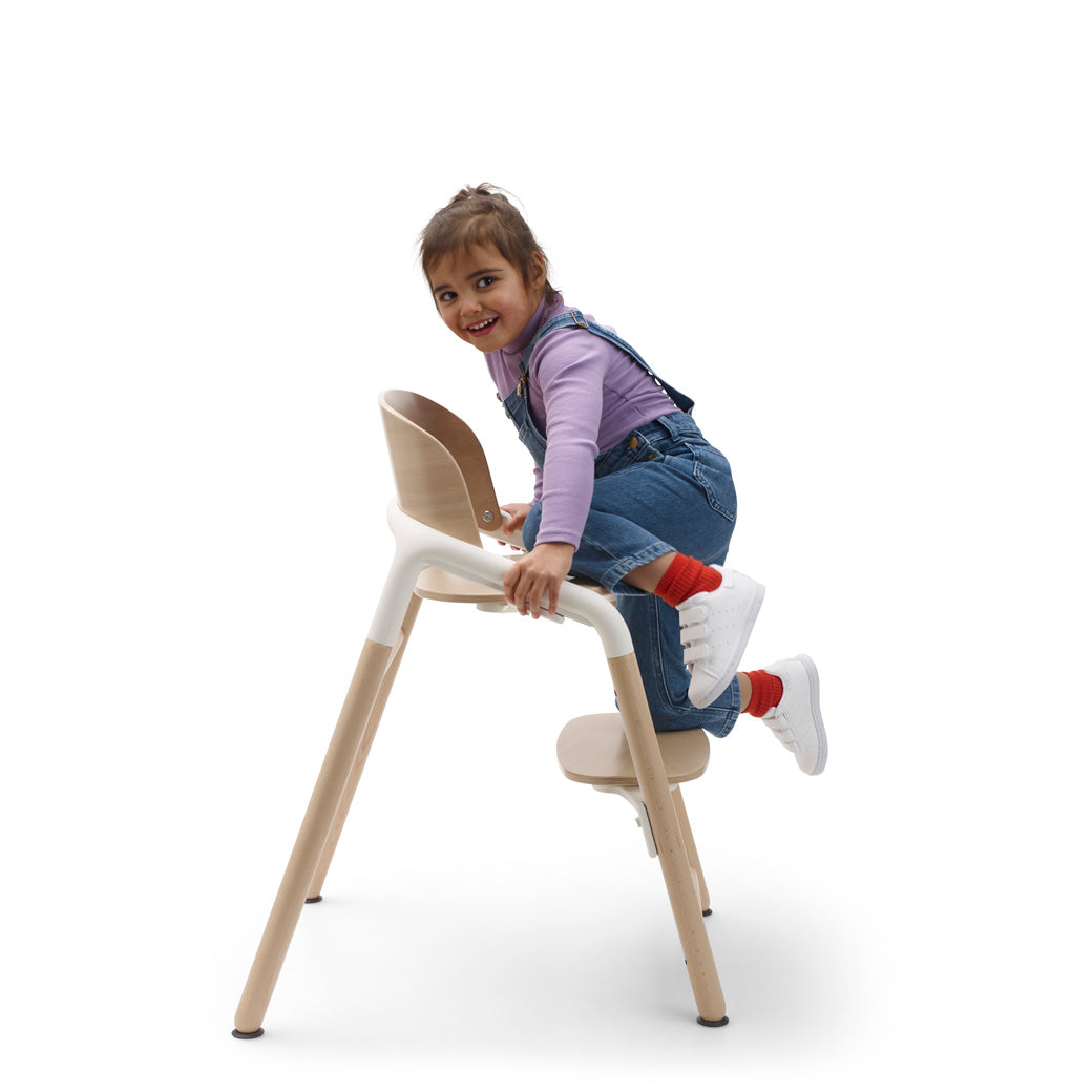 Side view of child climbing onto the Bugaboo Giraffe High Chair in --Color_Neutral Wood / White