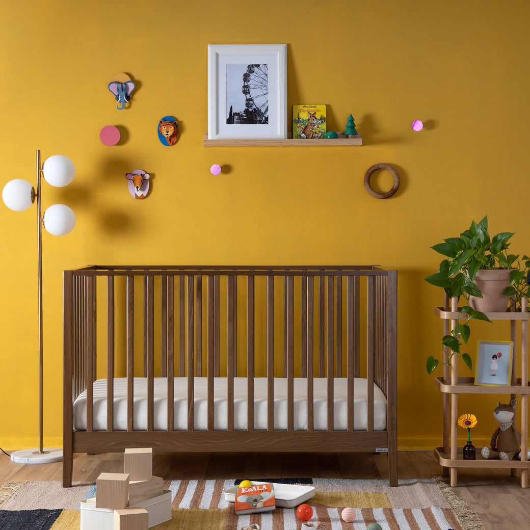 Front view of the Dadada Lala 3-in-1 Convertible Crib in a yellow room with toys  in - Color_Walnut