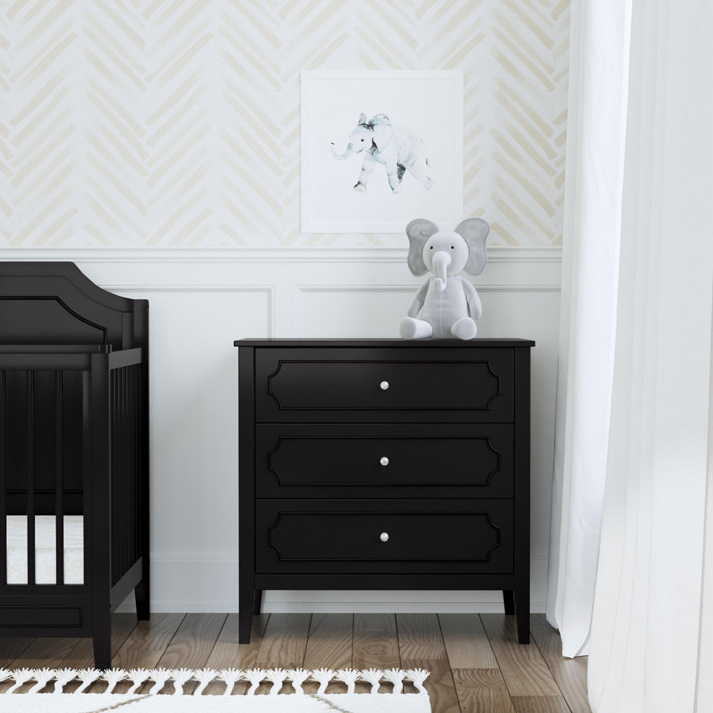 Front view of DaVinci Chloe Regency 3-Drawer Dresser next to a crib  in -- Color_Ebony