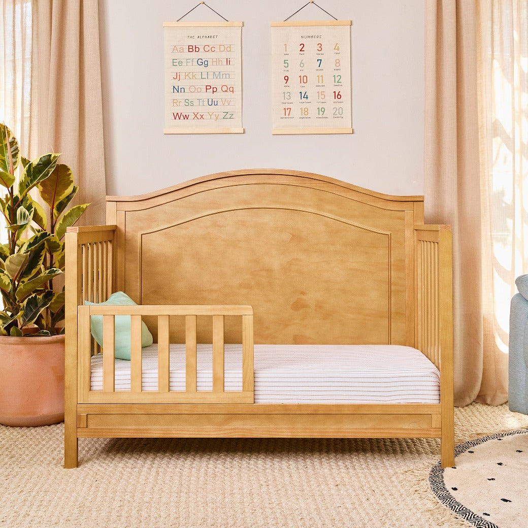 The DaVinci Charlie 4-in-1 Convertible Crib as toddler bed next to plant  in -- Color_Honey