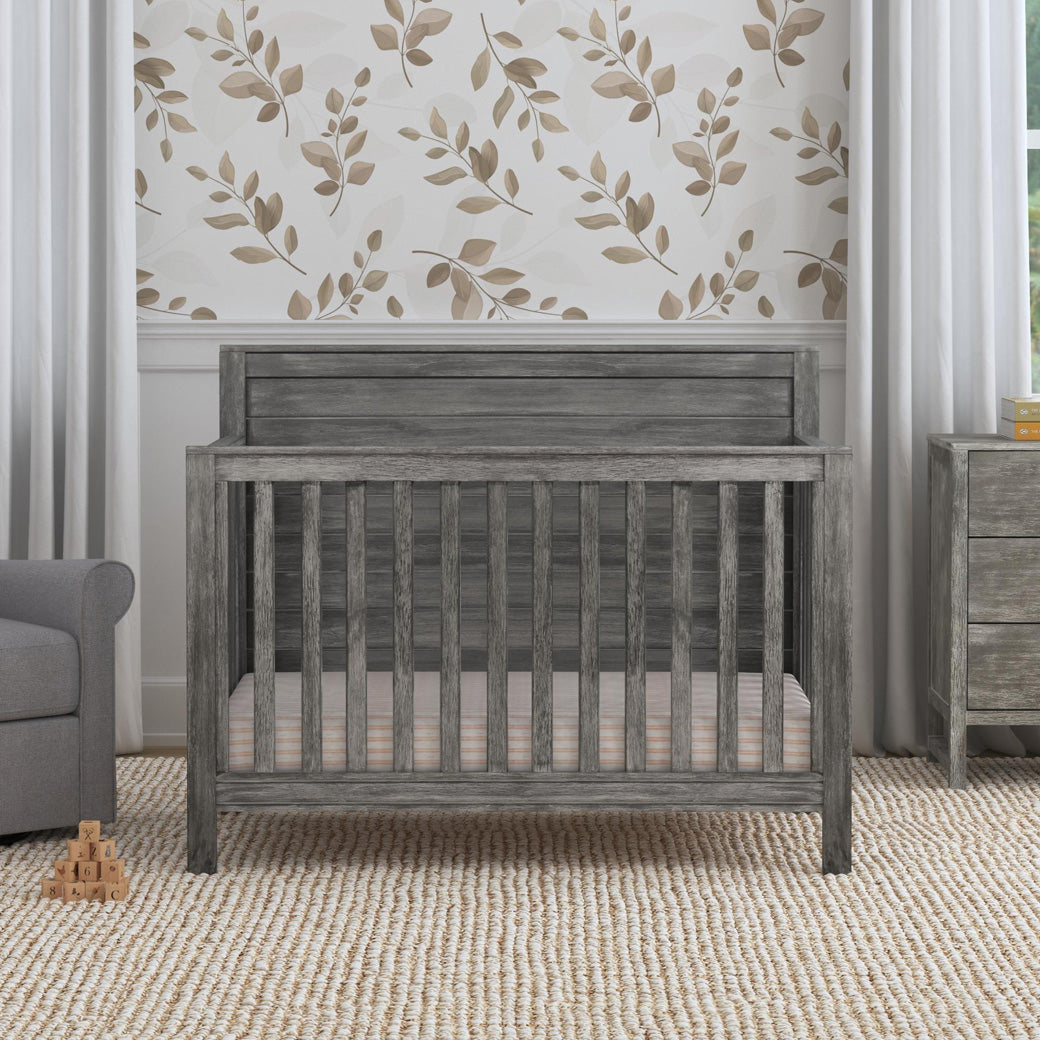 Front view of DaVinci Fairway 4-in-1 Convertible Crib next to a recliner and dresser  in -- Color_Cottage Grey