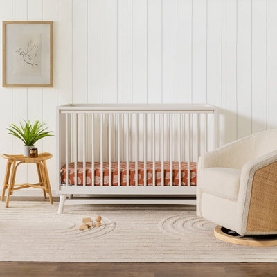 Babyletto's Peggy 3-in-1 Convertible Crib next to a table and recliner  in -- Color_Warm White