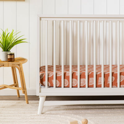 Front view of Babyletto's Peggy 3-in-1 Convertible Crib next to a table with a plant  in -- Color_Warm White
