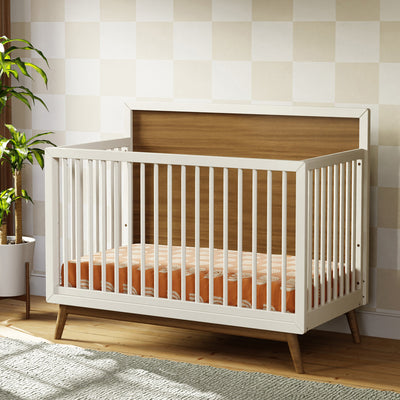 Babyletto's Palma 4-in-1 Convertible Crib next to a plant in -- Color_Warm White with Natural Walnut