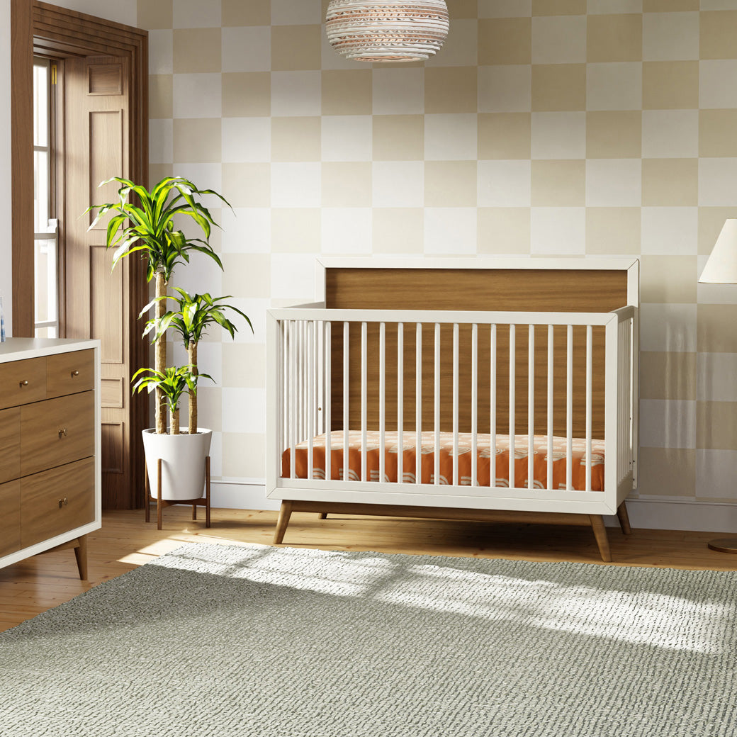 Babyletto's Palma 4-in-1 Convertible Crib next to a plant and dresser  in -- Color_Warm White with Natural Walnut