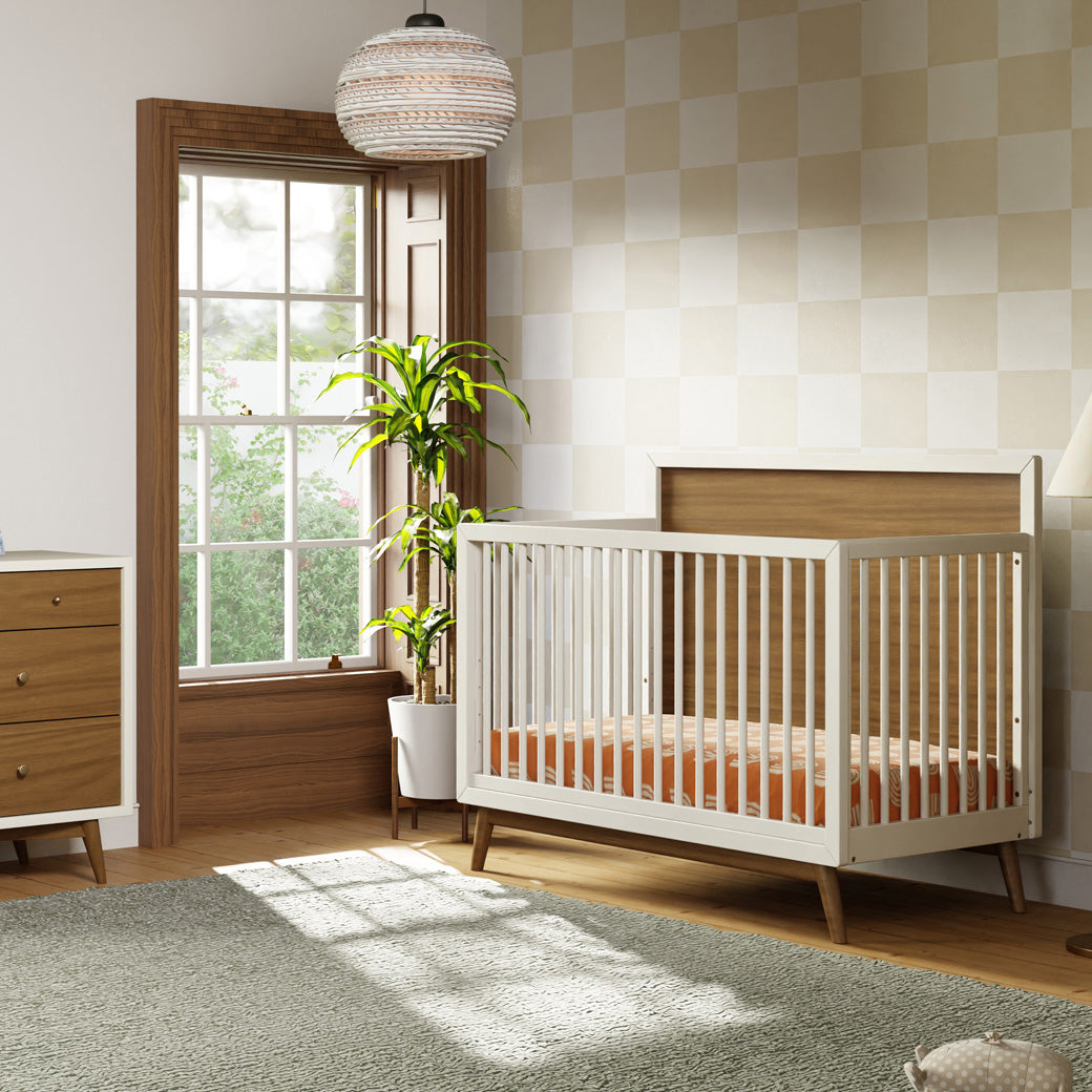 Babyletto's Palma 4-in-1 Convertible Crib next to a window and plant  in -- Color_Warm White with Natural Walnut