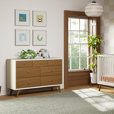 Babyletto's Palma 7-Drawer Assembled Double Dresser next to a window and crib  in -- Color_Warm White with Natural Walnut