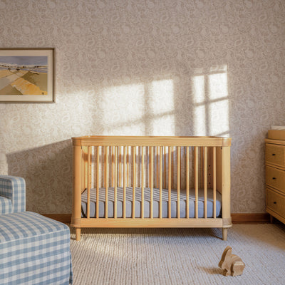 Front view of The Namesake Marin 3-in-1 Convertible Crib next to recliner and dresser in -- Color_Honey/Honey Cane