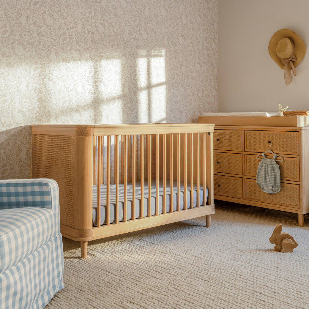 The Namesake Marin 3-in-1 Convertible Crib next to recliner and dresser in -- Color_Honey/Honey Cane