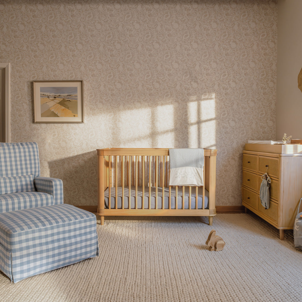 The Namesake Marin 3-in-1 Convertible Crib next to recliner, ottoman, and dresser  in -- Color_Honey/Honey Cane