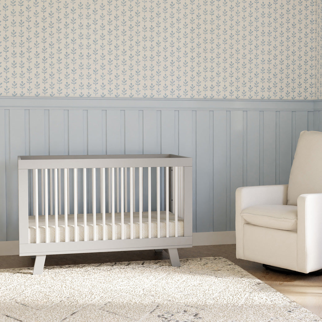 Babyletto Hudson 3-in-1 Convertible Crib + Toddler Rail next to a white recliner  in -- Color_White/Grey