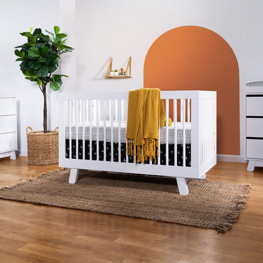 Babyletto Hudson 3-in-1 Crib next to a plant with a yellow blanket over the rail  in -- Color_White