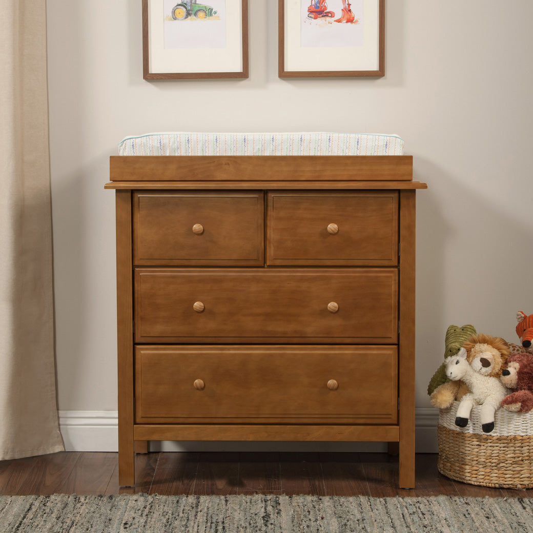 Front view of DaVinci Autumn 4-Drawer Dresser with changing tray  next to a basket with toys in -- Color_Chestnut