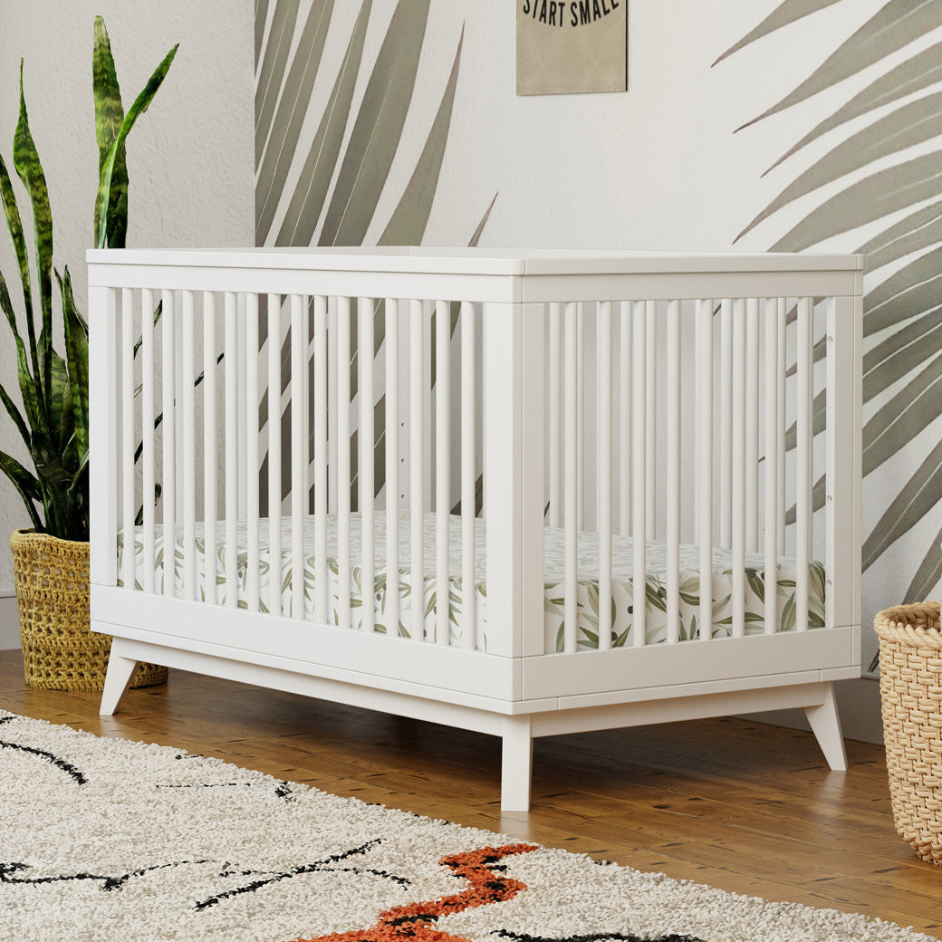 Babyletto's Scoot 3-in-1 Convertible Crib next to a plant and basket  in -- Color_White