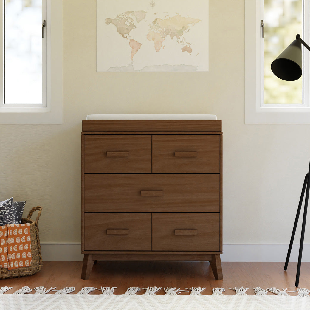 Front view of Babyletto's Scoot 3-Drawer Changer Dresser next to a basket and lamp in -- Color_Natural Walnut