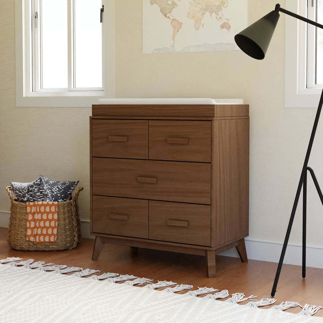 Babyletto's Scoot 3-Drawer Changer Dresser next to a basket  in -- Color_Natural Walnut