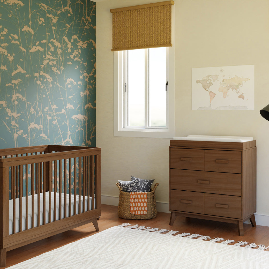 Babyletto's Scoot 3-Drawer Changer Dresser next to a basket and crib in -- Color_Natural Walnut