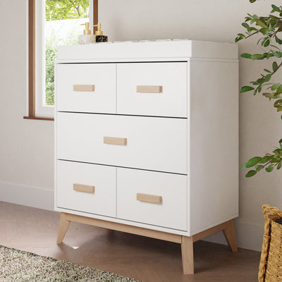 Babyletto's Scoot 3-Drawer Changer Dresser next to a window in -- Color_Washed Natural/White