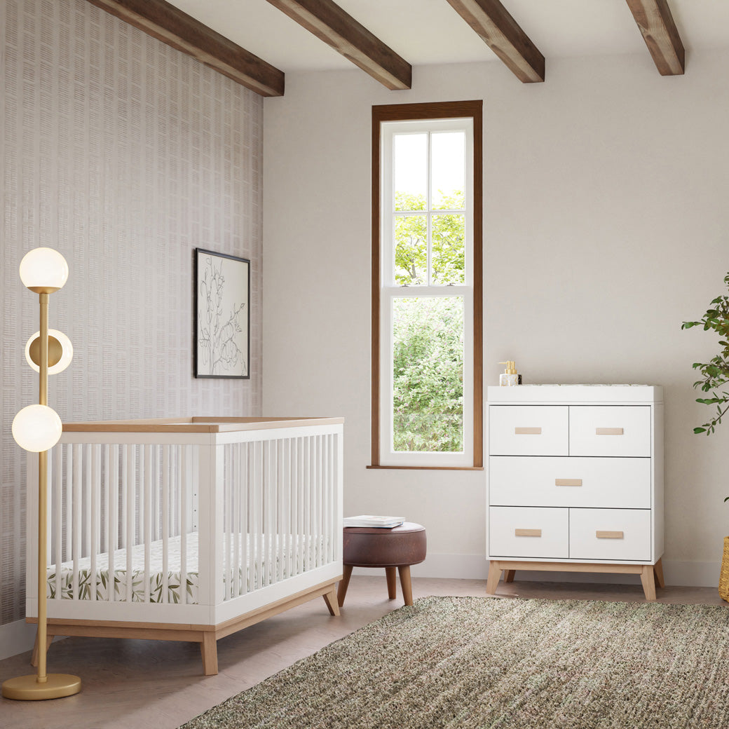 Babyletto's Scoot 3-Drawer Changer Dresser next to a window, stool, and crib  in -- Color_Washed Natural/White