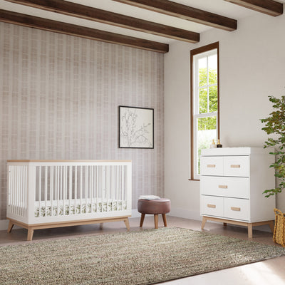 Babyletto's Scoot 3-Drawer Changer Dresser next to a window and crib  in -- Color_Washed Natural/White