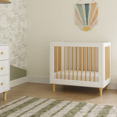 The Babyletto Lolly 4-in-1 Convertible Mini Crib next to a dresser in -- Color_White / Natural