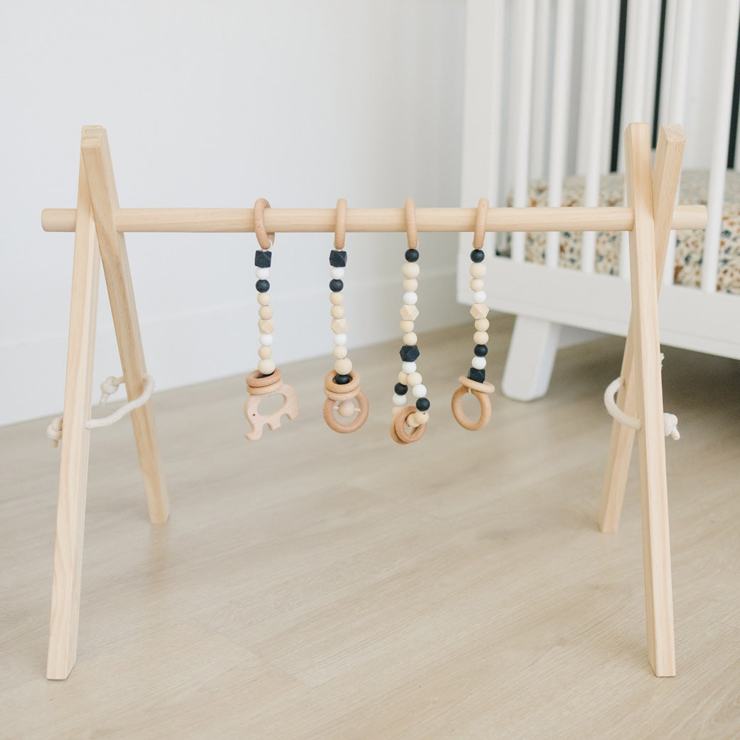 Poppyseed Play Wood Baby Gym + Toys in front of a white crib in -- Color_Natural _ Black