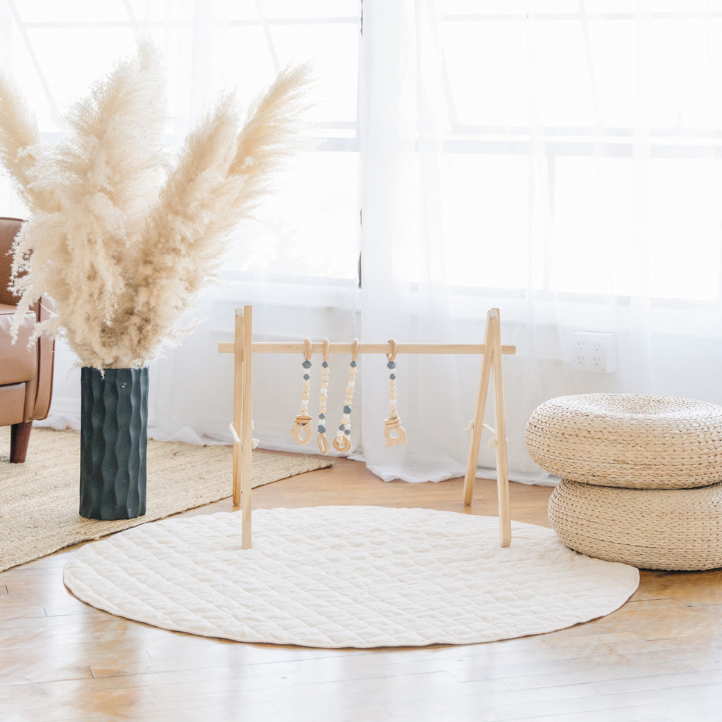 Poppyseed Play Wood Baby Gym + Toys on a white mat next to a vase and floor poufs in -- Color_Natural _ Gray