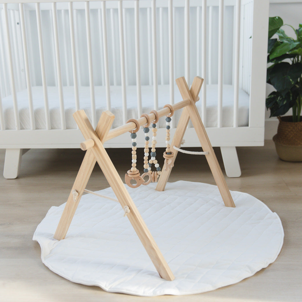 Poppyseed Play Wood Baby Gym + Toys on a mat in front of a white crib in -- Color_Natural _ Gray
