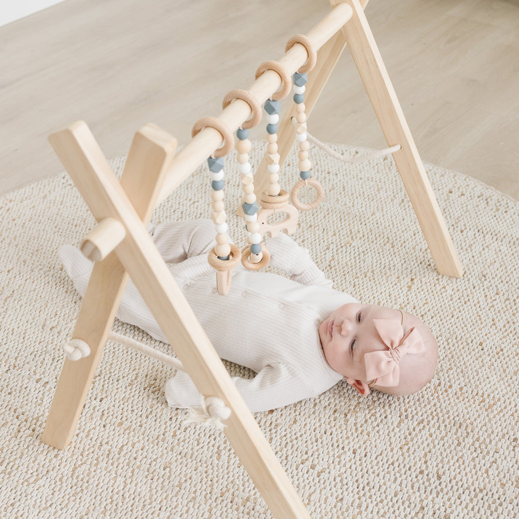 Baby laying and playing with the Poppyseed Play Wood Baby Gym + Toys in -- Color_Natural _ Gray